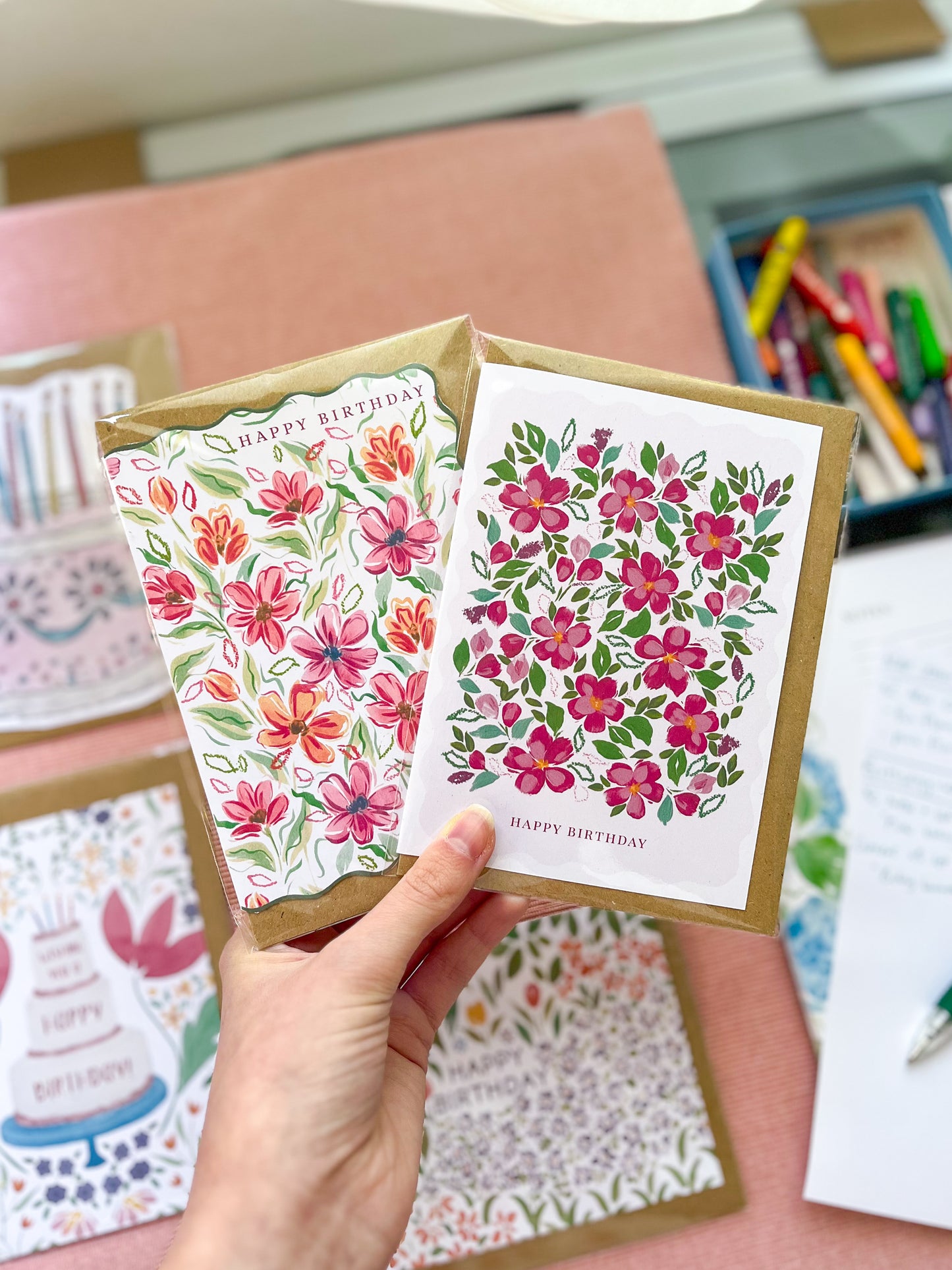 Hand holding two floral birthday cards with a blurred background of books and stationery.