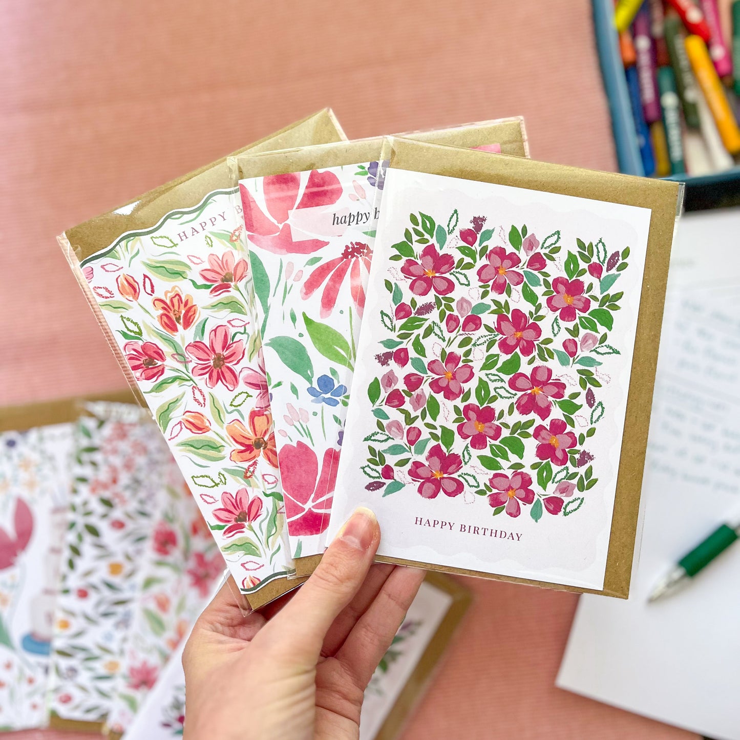 Hand holding three floral greeting cards with a pink surface and stationery items in the background.