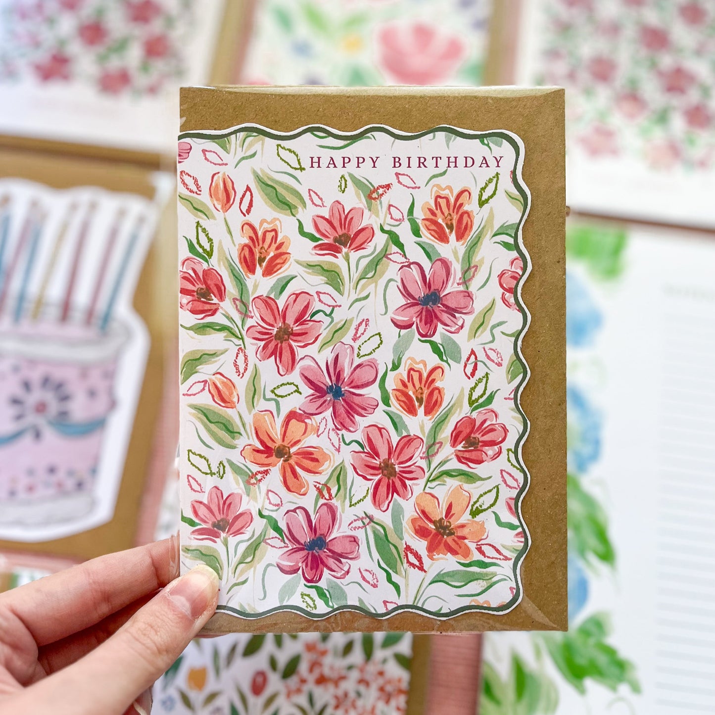 Handheld birthday card with floral design and 'Happy Birthday' text, surrounded by more cards in the background.