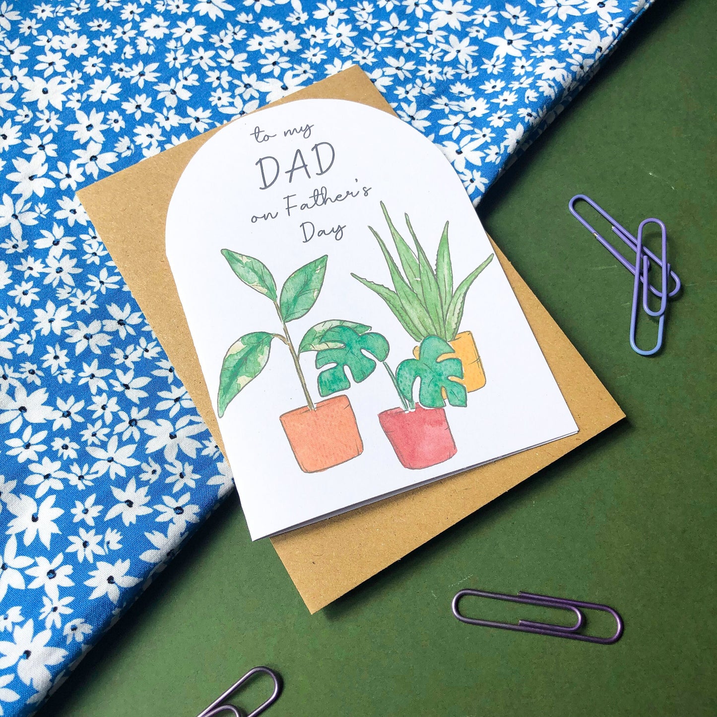 A Father's Day card displaying the words "DAD Happy Father's Day" above a collection of several house plants, each growing in a different coloured pot. The rich hues of the pots range from earthy green to bright orange and deep blue, complementing perfectly the vibrant green leaves of the plants. The card is an attractive and personal way to convey warm greetings and well wishes to a cherished dad on his special day.