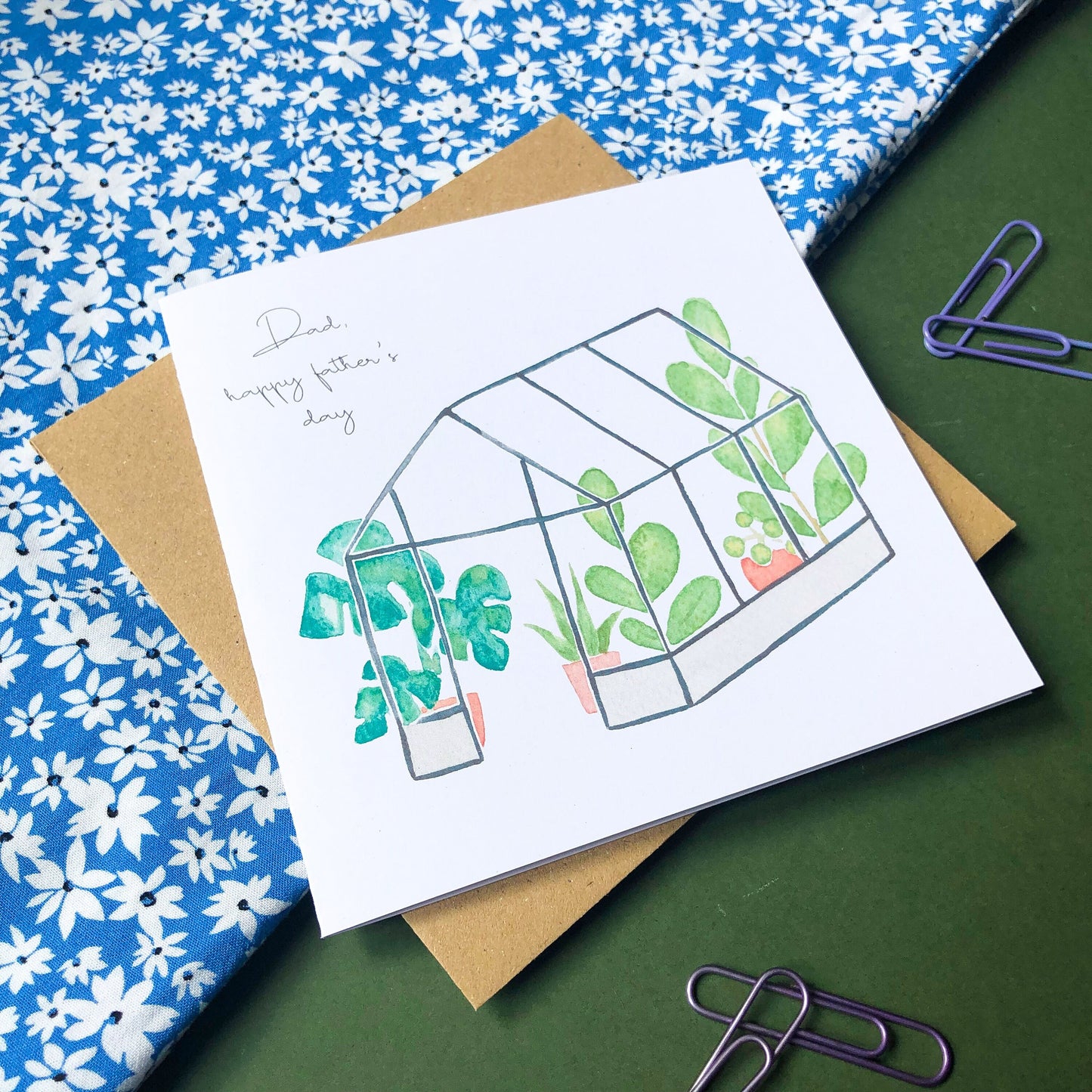 A square Father's Day card with a beautifully painted image of a tropical greenhouse, teeming with lush foliage and exotic blooms. The card also features the words "Dad Happy Father's Day" in an elegant cursive font, written in a contrasting colourful tone to the background painting. This card is a thoughtful and striking way to honour a special dad on his day, with the vibrant colours and intricate details of the painting adding to its unique appeal.