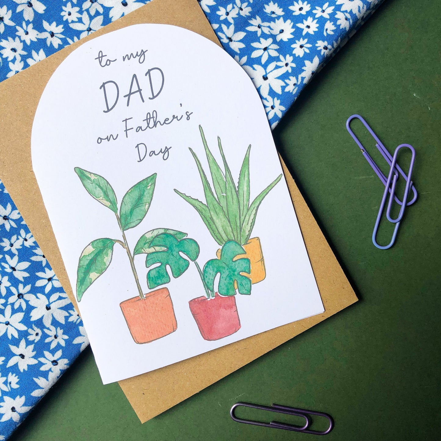 A Father's Day card displaying the words "DAD Happy Father's Day" above a collection of several house plants, each growing in a different coloured pot. The rich hues of the pots range from earthy green to bright orange and deep blue, complementing perfectly the vibrant green leaves of the plants. The card is an attractive and personal way to convey warm greetings and well wishes to a cherished dad on his special day.