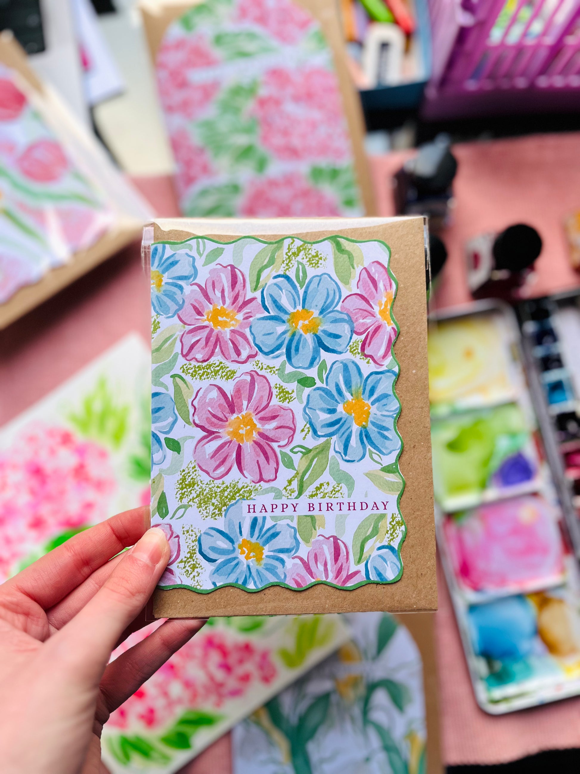 Floral-themed card with 'Happy Birthday' text held by a hand, surrounded by more cards in the background.