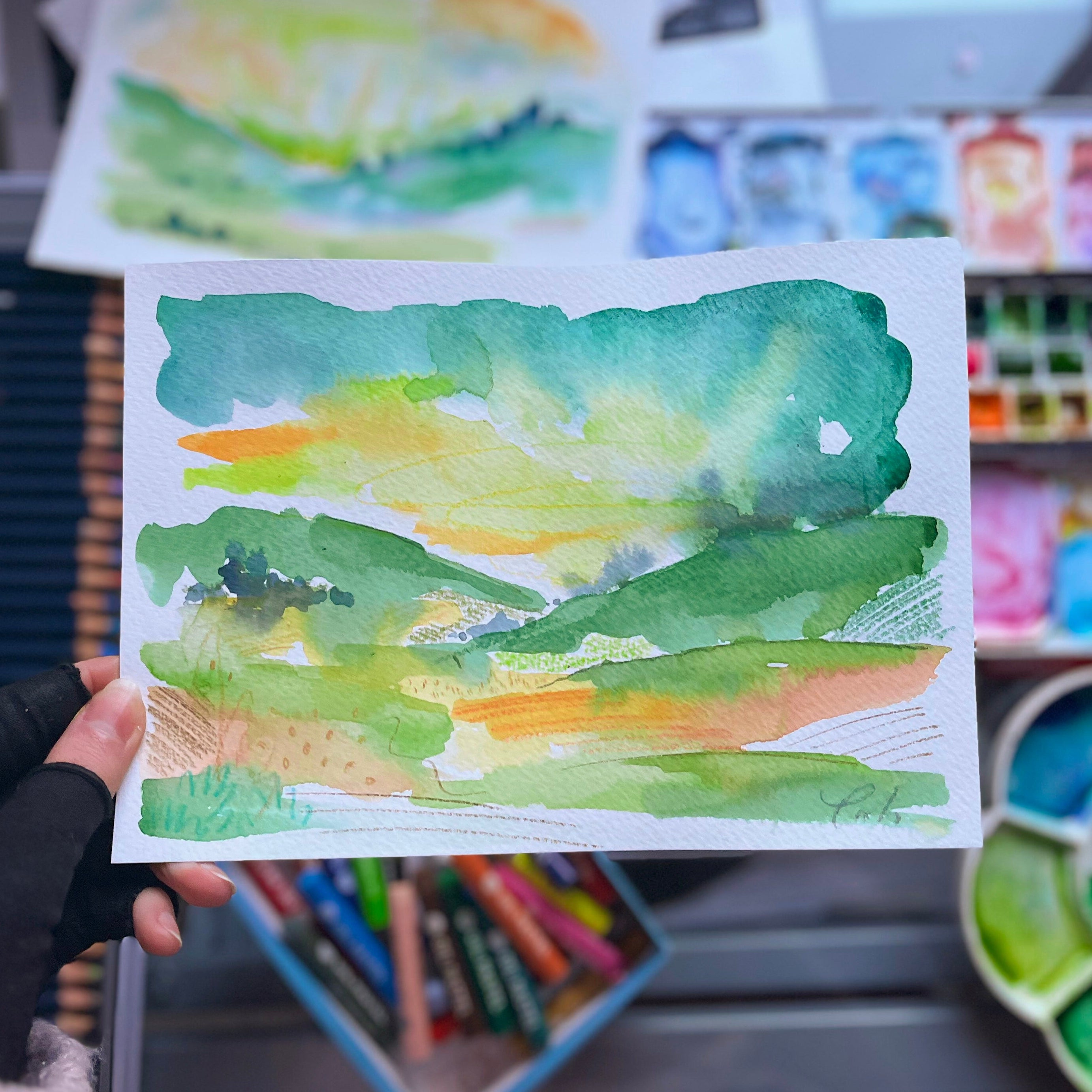Hand holding a watercolour painting of green and orange abstract landscape with art supplies in the background.