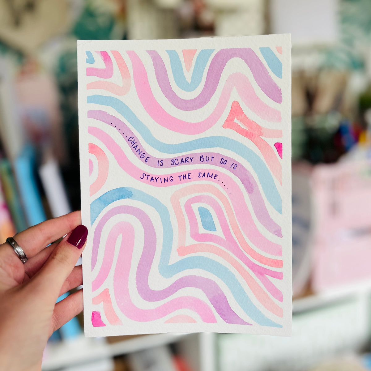 A hand with dark red nail polish holds a white art print featuring pastel pink, purple, and blue wavy lines with the text "CHANGE IS SCARY BUT SO IS STAYING THE SAME."