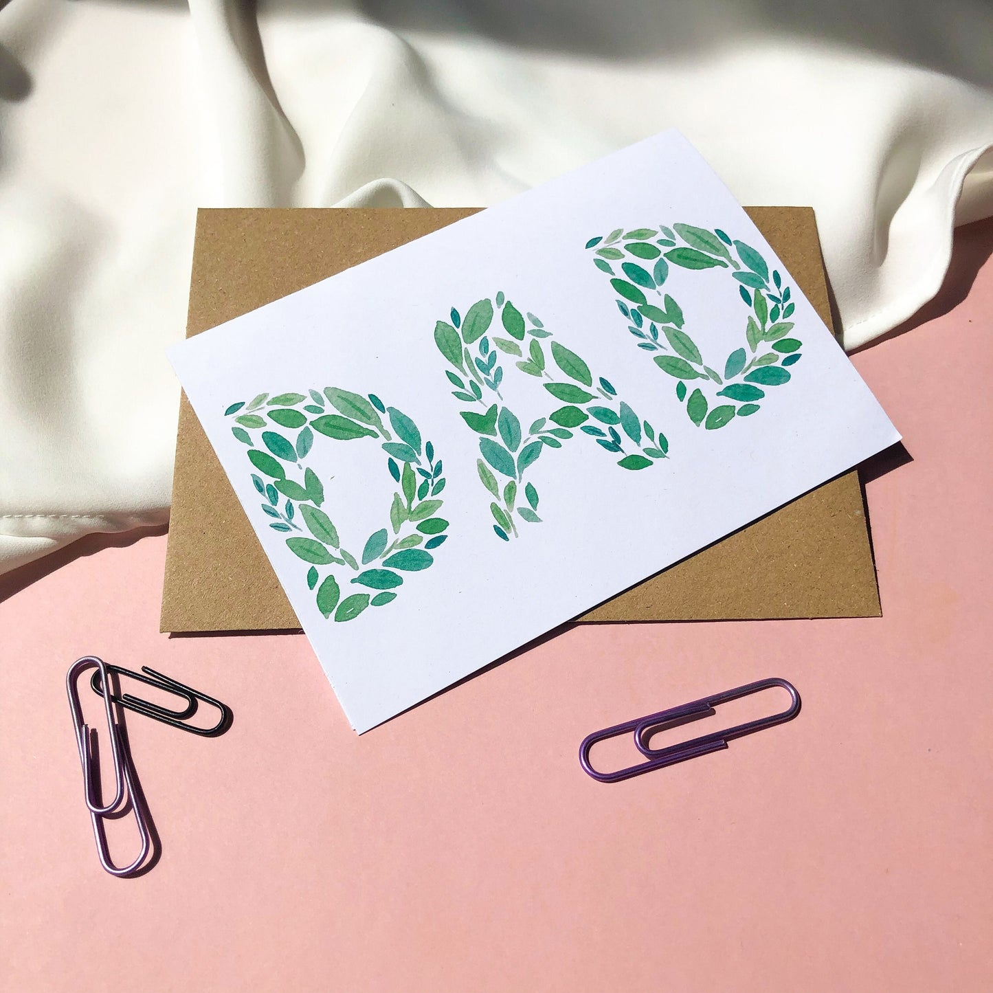 A Father's Day card with the word "DAD" spelled out beautifully in green leaves of various shades and shapes. The card offers a simple yet elegant way to show appreciation and affection for a beloved dad on his special day. the card is being held against a pink background