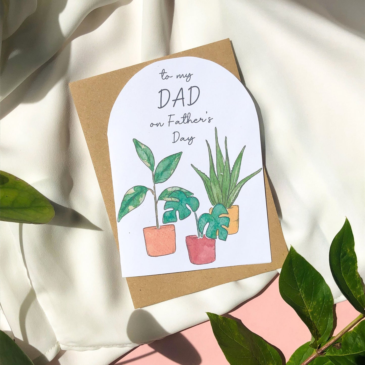 A Father's Day card displaying the words "DAD Happy Father's Day" above a collection of several house plants, each growing in a different coloured pot. The rich hues of the pots range from earthy green to bright orange and deep blue, complementing perfectly the vibrant green leaves of the plants. The card is an attractive and personal way to convey warm greetings and well wishes to a cherished dad on his special day.