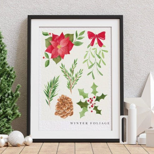A framed winter foliage art print featuring red poinsettias, green leaves, a pinecone, and holly on a white background, styled in a home setting with a small Christmas tree and holiday ornaments.